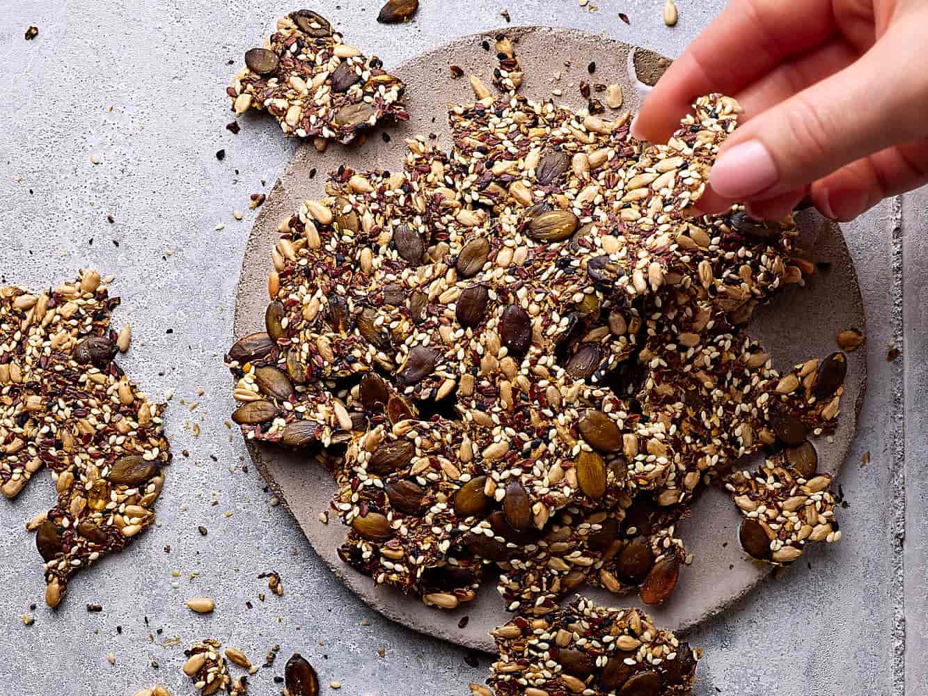 Saatencracker ohne Mehl auf einer Steingutplatte mit Körner-Crackern drumherum. Eine Hand greift nach einem Cracker.