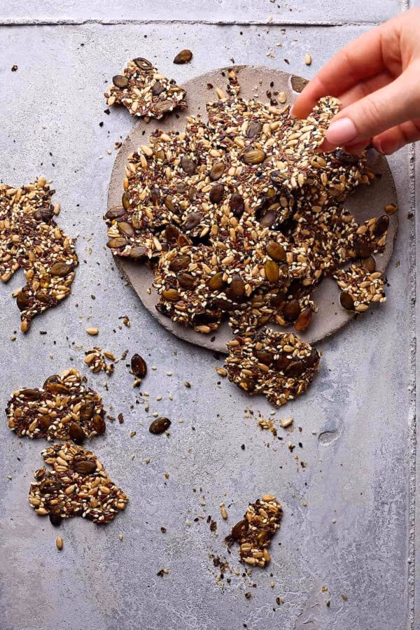 Saatencracker ohne Mehl auf einer Steingutplatte mit Körner-Crackern drumherum. Eine Hand greift nach einem Cracker.