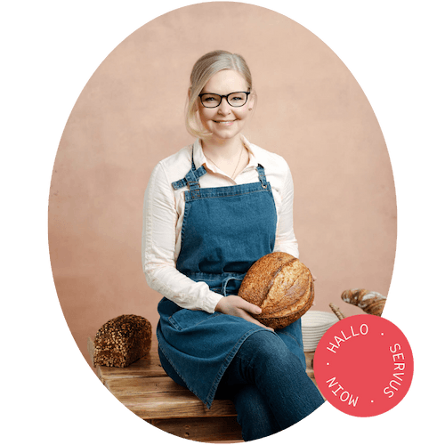 Sonja Bauer von Cookie und Co auf einem Holztisch sitzend meit einem Brot in der Hand und einem Glas Sauerteig daneben sowie weiteren Broten im Hintergrund.