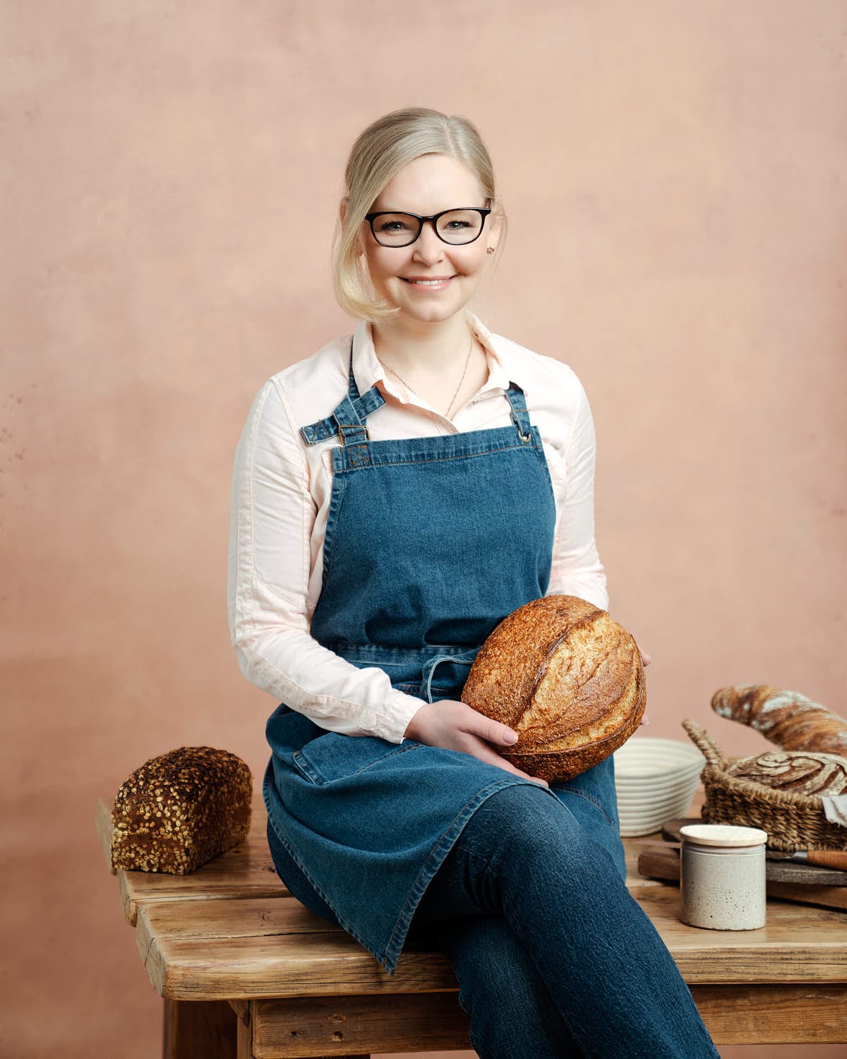 Sonja Bauer von Cookie und Co auf einem Holztisch sitzend meit einem Brot in der Hand und einem Glas Sauerteig daneben sowie weiteren Broten im Hintergrund.