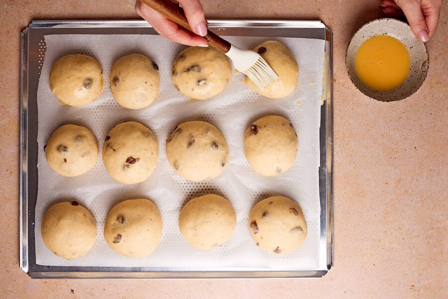 Reife Rosinebrötchen-Teiglinge auf einem Backblech, die vor dem backen mit Eistreiche eingepinselt werden.
