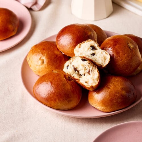 Rosinenbrötchen auf einem rosa Teller, eines aufgebrochen mit weicher Krume und sichtbaren Rosinen, im Frühstücks-Setting.