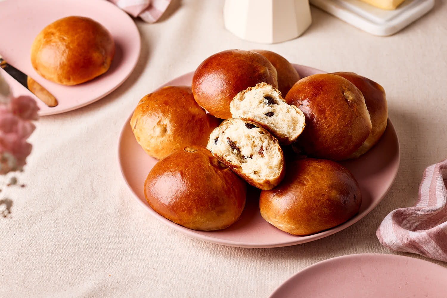 Rosinenbrötchen auf einem rosa Teller, eines aufgebrochen mit weicher Krume und sichtbaren Rosinen, im Frühstücks-Setting.