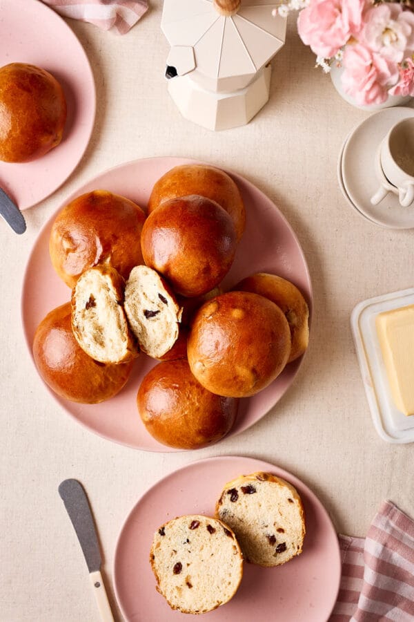Rosinenbrötchen auf einem rosa Teller, eines aufgeschnitten mit saftiger Krume und prallen Rosinen, daneben Butter und eine Tasse Kaffee.