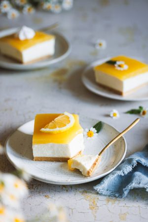 Lemon Cheesecake, ein fruchtiger Zitronen-Käsekuchen auf einem Teller mit einer Kuchengabel und weiteren Kuchenstücken im Hintergrund.