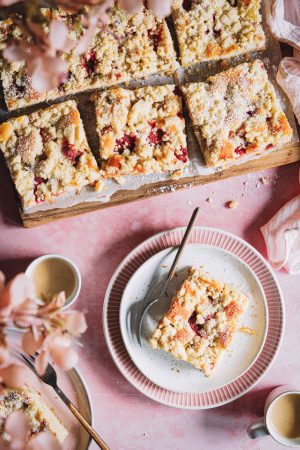 Ein Stück Erdbeer-Rhabarber-Kuchen auf einem Teller mit zwei Tassen Kaffee daneben und einem Brett mit weiteren Kuchenstücken im Hintergrund.